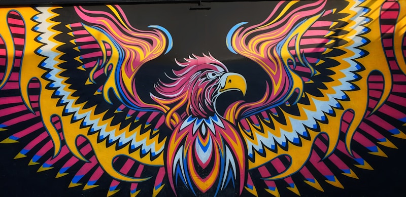 A vibrant, abstract mural featuring a stylized eagle with outstretched wings. The artwork showcases intricate patterns with zig-zag and curved lines, using bright colors such as pink, yellow, blue, and black. The eagle's eyes and beak are emphasized, giving it a fierce and dynamic appearance.