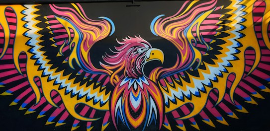 A vibrant, abstract mural featuring a stylized eagle with outstretched wings. The artwork showcases intricate patterns with zig-zag and curved lines, using bright colors such as pink, yellow, blue, and black. The eagle's eyes and beak are emphasized, giving it a fierce and dynamic appearance.