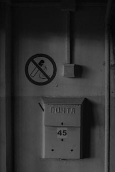A grayscale image features an old metal mailbox mounted on a wall, with the word 'ПОЧТА' embossed on it and the number '45' displayed below. Above the mailbox, a sign depicts a cigarette with a diagonal line through it, indicating no smoking is allowed. The wall appears weathered and has electrical conduits running vertically and horizontally nearby.