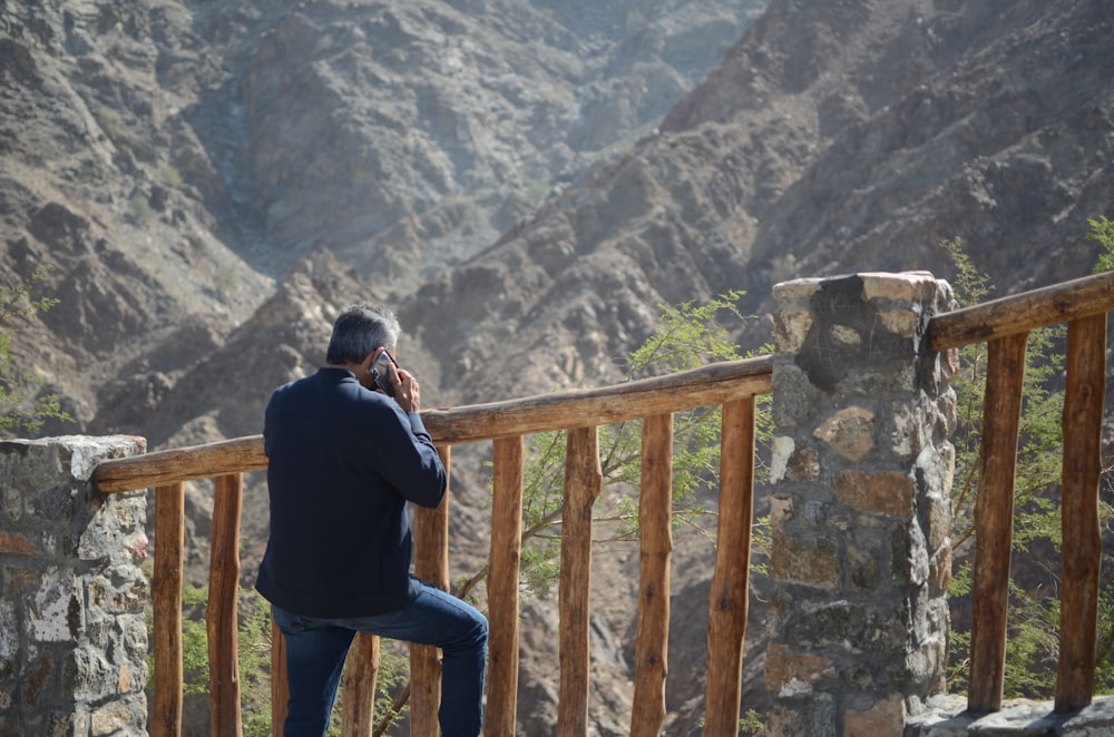 man in black jacket and blue denim jeans sitting on brown wooden fence during daytime