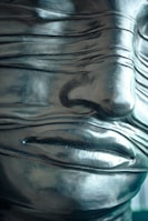 A close-up of a sculptural face with a smooth, metallic finish, featuring a series of narrow, horizontal bands wrapping across the surface. The bands cover the eyes, adding an enigmatic quality to the artwork. The material appears polished, reflecting light subtly.