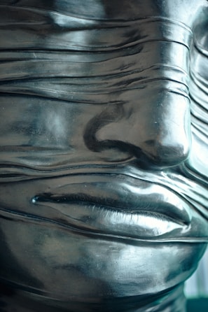 A close-up of a sculptural face with a smooth, metallic finish, featuring a series of narrow, horizontal bands wrapping across the surface. The bands cover the eyes, adding an enigmatic quality to the artwork. The material appears polished, reflecting light subtly.