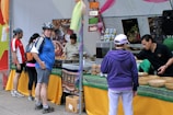 A busy market stall with several people interacting. A man wearing a helmet and a blue jersey stands near the front, looking at the camera. Various foods and drinks are set out on the table, with signs indicating different flavors. Staff are assisting customers with their selections.