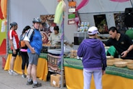 A busy market stall with several people interacting. A man wearing a helmet and a blue jersey stands near the front, looking at the camera. Various foods and drinks are set out on the table, with signs indicating different flavors. Staff are assisting customers with their selections.
