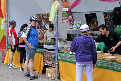 A busy market stall with several people interacting. A man wearing a helmet and a blue jersey stands near the front, looking at the camera. Various foods and drinks are set out on the table, with signs indicating different flavors. Staff are assisting customers with their selections.