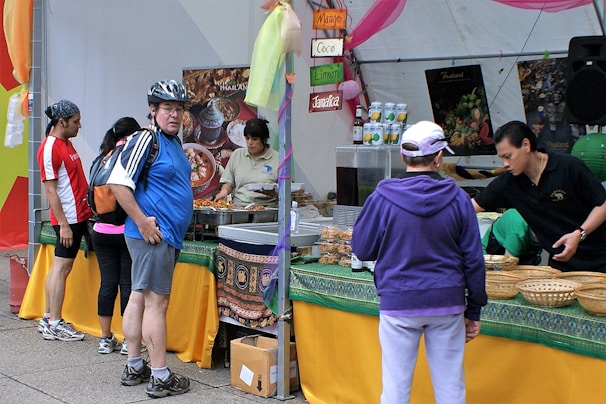 A busy market stall with several people interacting. A man wearing a helmet and a blue jersey stands near the front, looking at the camera. Various foods and drinks are set out on the table, with signs indicating different flavors. Staff are assisting customers with their selections.