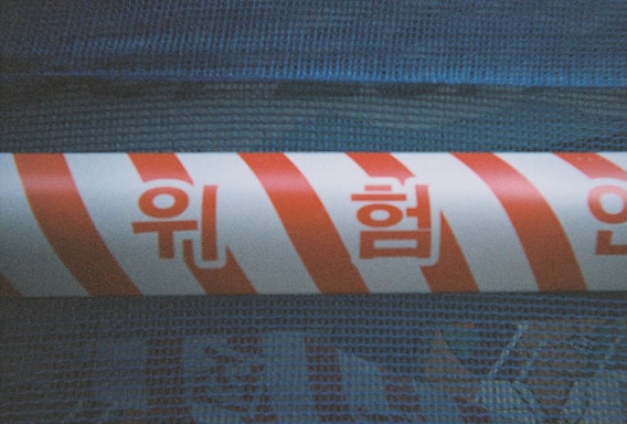 A section of a barrier tape with red and white diagonal stripes is wrapped around a textured blue surface. Korean characters are printed in red across the tape.