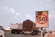 A traffic scene with a prominent speed limit sign displaying 50 in the foreground. Several large trucks and vehicles are visible on the road, moving in the same direction. The sky is partly cloudy with a soft color palette.