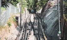 The image features a funicular railway track set in a natural landscape. The tracks run through a narrow passage with steeply inclined surroundings covered in greenery and rocky surfaces. Lamp posts line the track on both sides, and a cable car is visible in the distance at the top of the hill. Metal railings and a mesh fence are present on the left side.