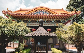 An ornate, traditional Asian temple features intricate architectural details, including a brightly colored roof and decorative carvings. Red lanterns hang prominently, and the temple is surrounded by lush greenery. A small pavilion with a dark, pagoda-style roof is situated in the foreground.