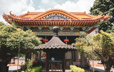 An ornate, traditional Asian temple features intricate architectural details, including a brightly colored roof and decorative carvings. Red lanterns hang prominently, and the temple is surrounded by lush greenery. A small pavilion with a dark, pagoda-style roof is situated in the foreground.