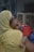 A woman wearing a yellow garment is holding a small child. The child, dressed in a red and blue outfit, rests contentedly on the woman's shoulder. The setting appears to be indoors with a neutral background, and the scene conveys a tender moment between the two.