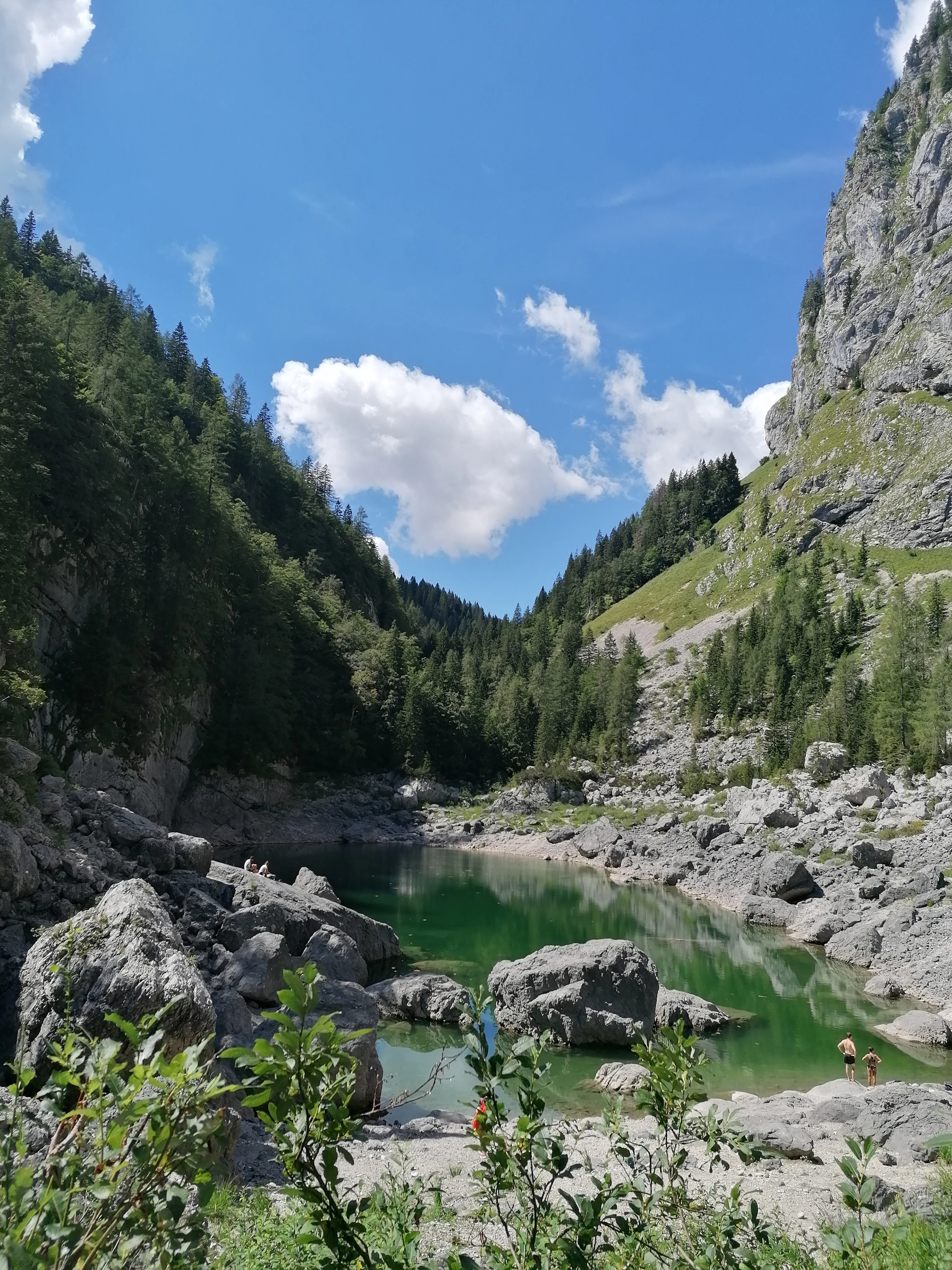 A green mountain lake among boulders and spruce beneath limestone crags, with small figures at the shore