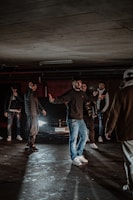 A group of people standing in a dimly lit parking garage in front of a car with headlights on. The individuals are casually dressed, mostly in dark clothing and jeans. The atmosphere is moody with low lighting and shadows cast on the concrete floor.