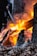 A vibrant and intense fire is consuming pieces of wood, with bright orange and yellow flames reaching upwards. Charring and ashes are visible on the wooden surfaces. There are fallen dried leaves around the fire, and someone in dark clothing is tending to the fire with a tool.