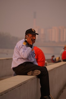 A man in a uniform is sitting on a concrete ledge, engaged in a phone call. He has a cap on and appears to be in an urban setting with towers and buildings slightly blurred in the background.