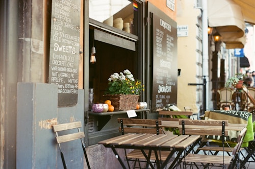A charming outdoor cafe scene with wooden tables and chairs lined up along a narrow street. The chalkboard menu displays a list of beverages and sweets offered by the cafe. A basket filled with flowers and small pumpkins is placed on a counter by the window. The overall atmosphere is cozy and inviting, with warm lighting and potted plants enhancing the ambiance.
