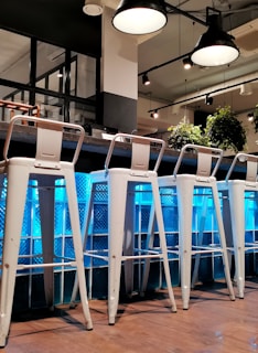 A modern bar setting with several tall white metal stools arranged in a row against a counter. The background features a textured blue glass panel that reflects light, creating an ambient atmosphere. Above, industrial-style pendant lights hang from the ceiling, adding to the contemporary design. Green plants add a touch of nature and contrast with the otherwise sleek and minimalistic interior.