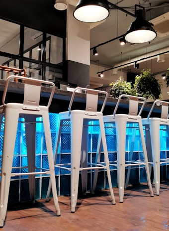 A modern bar setting with several tall white metal stools arranged in a row against a counter. The background features a textured blue glass panel that reflects light, creating an ambient atmosphere. Above, industrial-style pendant lights hang from the ceiling, adding to the contemporary design. Green plants add a touch of nature and contrast with the otherwise sleek and minimalistic interior.