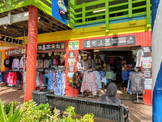 A colorful and vibrant storefront displays an array of clothing, including shirts and accessories, under a bright awning. The bright green, yellow, and blue color scheme dominates the building's exterior. Signage with catchy phrases and 'CBD Sold Here' are prominently displayed. A person with a hoodie sits on a bench in front of the shop, taking in the lively atmosphere.