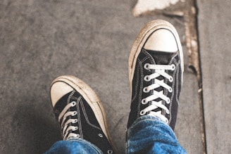 A person's feet wearing black canvas sneakers with white rubber caps and laces, sitting on a grey concrete surface. The jeans are slightly faded, giving a casual and relaxed appearance.