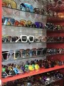 A collection of various sunglasses displayed on multiple glass shelves, featuring a range of colors and styles. The frames vary from modern and sleek to retro and bold designs, with lenses that are tinted in different hues such as blue, gold, and dark shades.