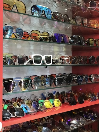 A collection of various sunglasses displayed on multiple glass shelves, featuring a range of colors and styles. The frames vary from modern and sleek to retro and bold designs, with lenses that are tinted in different hues such as blue, gold, and dark shades.