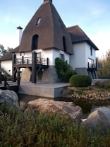 A unique house with a thatched, triangular roof and white walls is situated by a small pond. The building incorporates modern and rustic elements, with wooden balconies and stone foundations. The surrounding area features rocks, greenery, and shrubs, generating a tranquil and natural atmosphere.