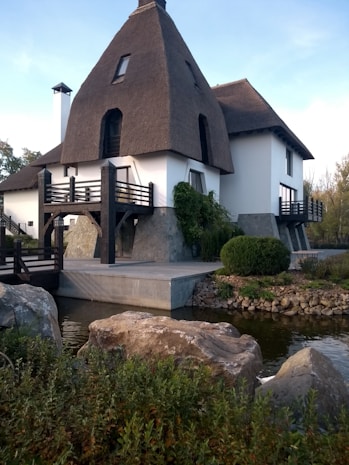 A unique house with a thatched, triangular roof and white walls is situated by a small pond. The building incorporates modern and rustic elements, with wooden balconies and stone foundations. The surrounding area features rocks, greenery, and shrubs, generating a tranquil and natural atmosphere.