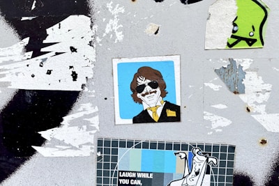 A surface with multiple overlapping stickers, including an illustration of a person with sunglasses and a yellow tie, a cartoonish green character, and a poster with the text 'LAUGH WHILE YOU CAN'. The background is a worn, grungy texture with remnants of torn paper.