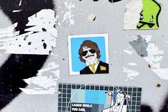 A surface with multiple overlapping stickers, including an illustration of a person with sunglasses and a yellow tie, a cartoonish green character, and a poster with the text 'LAUGH WHILE YOU CAN'. The background is a worn, grungy texture with remnants of torn paper.