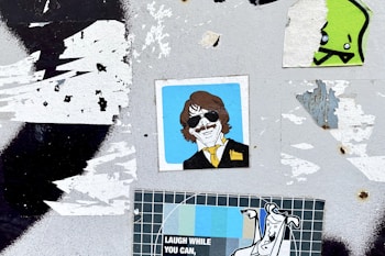 A surface with multiple overlapping stickers, including an illustration of a person with sunglasses and a yellow tie, a cartoonish green character, and a poster with the text 'LAUGH WHILE YOU CAN'. The background is a worn, grungy texture with remnants of torn paper.