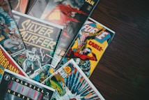 A collection of colorful comic book covers displayed on a wooden surface. The covers include titles like Silver Surfer, X-Force, and Spider-Man, featuring vibrant illustrations of superheroes and action scenes. Several comics are visible, with dynamic poses and dramatic artwork typical of comic book design.