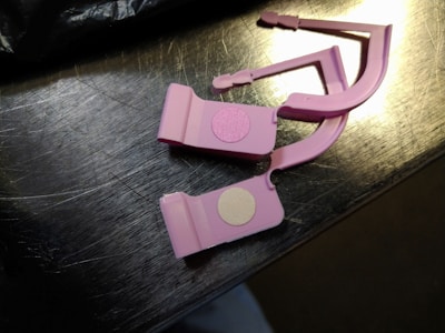 Two pink plastic clips with circular adhesive pads are placed on a metallic surface. The surface reflects light, showing a shiny appearance.