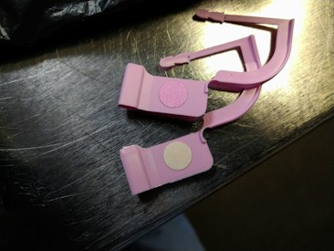 Two pink plastic clips with circular adhesive pads are placed on a metallic surface. The surface reflects light, showing a shiny appearance.