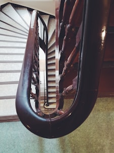 A descending spiral staircase with a curved wooden railing. The stairs are covered with light-colored steps, and the view looks downwards through the middle of the spiral, revealing additional levels below. The wall contrasts with the dark wood of the handrail, and the floor features green carpeting.