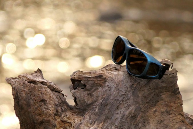 Wraparound sunglasses with brown lenses resting on a piece of driftwood, sunlight glittering on the water behind