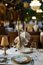 A sophisticated dining table setting with elegant folded napkins resembling birds, set in a well-lit, ambient dining area. Crystal wine glasses and cutlery are arranged neatly around white and gold-bordered plates. Warm light from chandeliers and lamps enhances the upscale atmosphere.