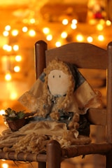 A handmade doll with curly yarn hair and a simple stitched face, sitting on a wooden chair. The doll is dressed in a blue and beige outfit, adorned with lace. To its left is a small woven basket holding a succulent plant. The background is warmly lit with soft, golden bokeh lights.