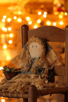 A handmade doll with curly yarn hair and a simple stitched face, sitting on a wooden chair. The doll is dressed in a blue and beige outfit, adorned with lace. To its left is a small woven basket holding a succulent plant. The background is warmly lit with soft, golden bokeh lights.