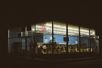 A laundromat is illuminated at night, visible from outside through large glass windows. The interior features rows of washing machines and dryers, and the walls are painted blue. An 'Open' neon sign is glowing inside.