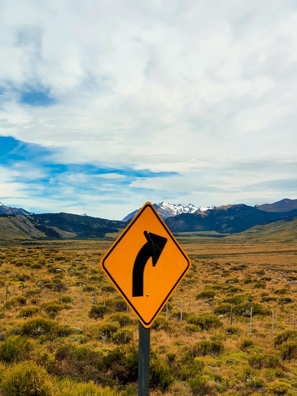 yellow and black road sign on green grass field under blue sky and white clouds during