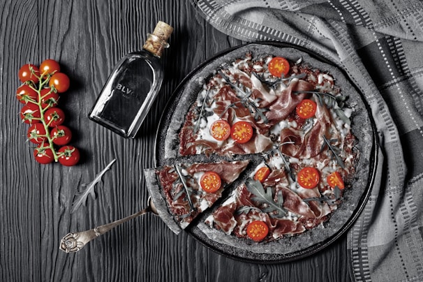 A gourmet pizza with a charcoal-black crust, topped with sliced cured meat, cherry tomato halves, and arugula, arranged on a dark, textured surface. A cluster of bright red cherry tomatoes on the vine lies beside a rustic silver serving utensil. A bottle labeled 'BLVK' with a cork lies next to the pizza, and a folded patterned cloth is draped nearby.