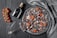 A gourmet pizza with a charcoal-black crust, topped with sliced cured meat, cherry tomato halves, and arugula, arranged on a dark, textured surface. A cluster of bright red cherry tomatoes on the vine lies beside a rustic silver serving utensil. A bottle labeled 'BLVK' with a cork lies next to the pizza, and a folded patterned cloth is draped nearby.