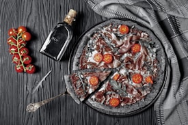 A gourmet pizza with a charcoal-black crust, topped with sliced cured meat, cherry tomato halves, and arugula, arranged on a dark, textured surface. A cluster of bright red cherry tomatoes on the vine lies beside a rustic silver serving utensil. A bottle labeled 'BLVK' with a cork lies next to the pizza, and a folded patterned cloth is draped nearby.