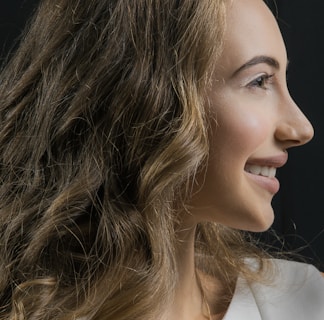A profile view of a woman with long, wavy brown hair and a smiling expression. The background is dark, highlighting her features and the texture of her hair. She is wearing a light-colored top.