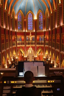 A grand, ornate cathedral interior with vibrant stained glass windows and intricate details. The ceiling is vaulted and decorated with arches and patterns, creating a majestic atmosphere. A person is seated at an organ in the foreground, focusing on music sheets, while a large illuminated crucifix hangs prominently in the background.