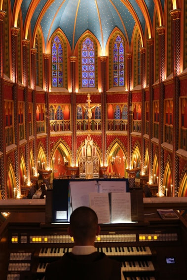 A grand, ornate cathedral interior with vibrant stained glass windows and intricate details. The ceiling is vaulted and decorated with arches and patterns, creating a majestic atmosphere. A person is seated at an organ in the foreground, focusing on music sheets, while a large illuminated crucifix hangs prominently in the background.