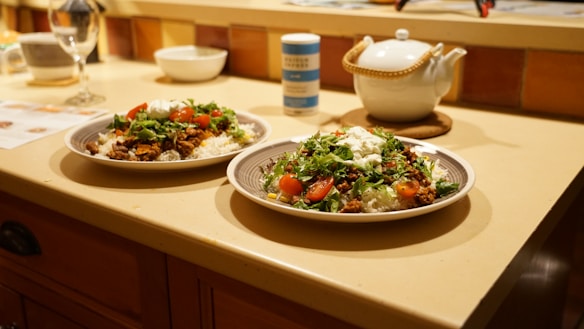Two plates of food are placed on a kitchen counter, featuring rice topped with ground meat, shredded lettuce, sliced cherry tomatoes, and a dollop of white sauce. A teapot with a wicker handle, a container of seasoning, and an empty wine glass are also present on the counter. The tiles and lighting give a warm ambiance.