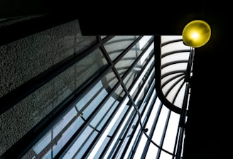 A glass roof structure featuring a series of curved, intersecting beams and transparent panels. A glowing light fixture in the upper right corner adds warmth to the otherwise cool-toned architectural elements, creating a dynamic play of lines and light.